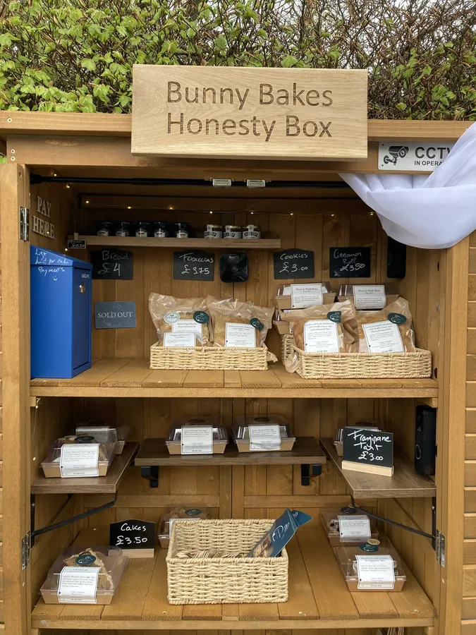 The Bunny Bakes honesty box, fully stocked: wooden shelves of wrapped bakes in baskets beneath a carved Bunny Bakes Honesty Box sign.