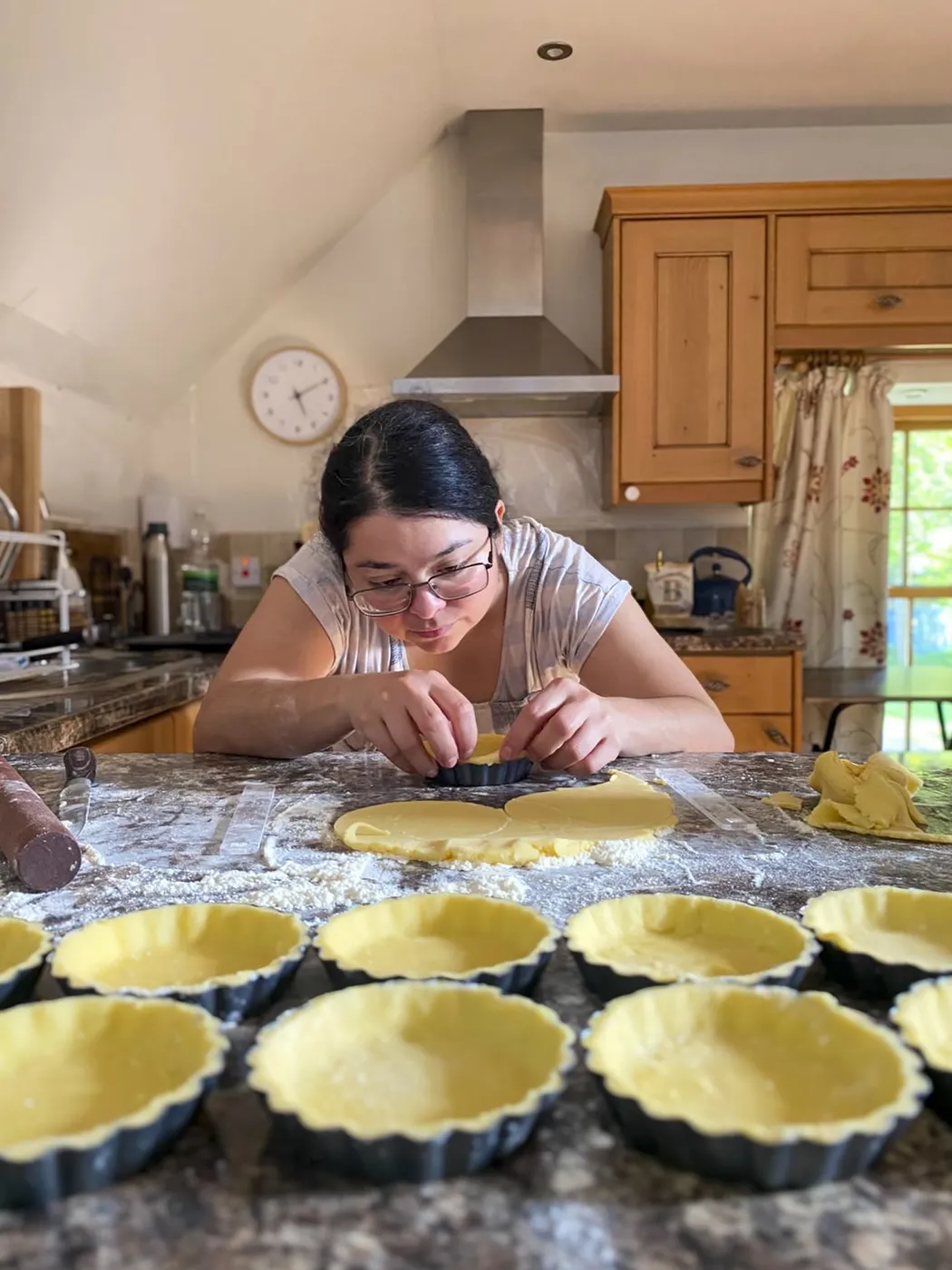 Maya crimping tart shells at a kitchen counter dusted with flour, a dozen tart tins lined up in front of her