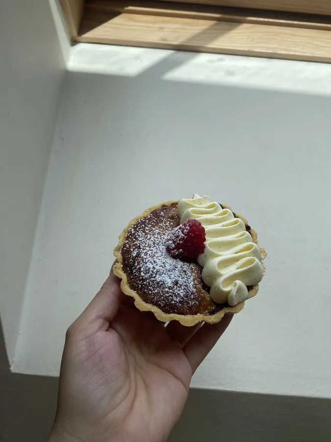 A hand holding a small tartlet topped with piped cream, a raspberry and a dusting of sugar.