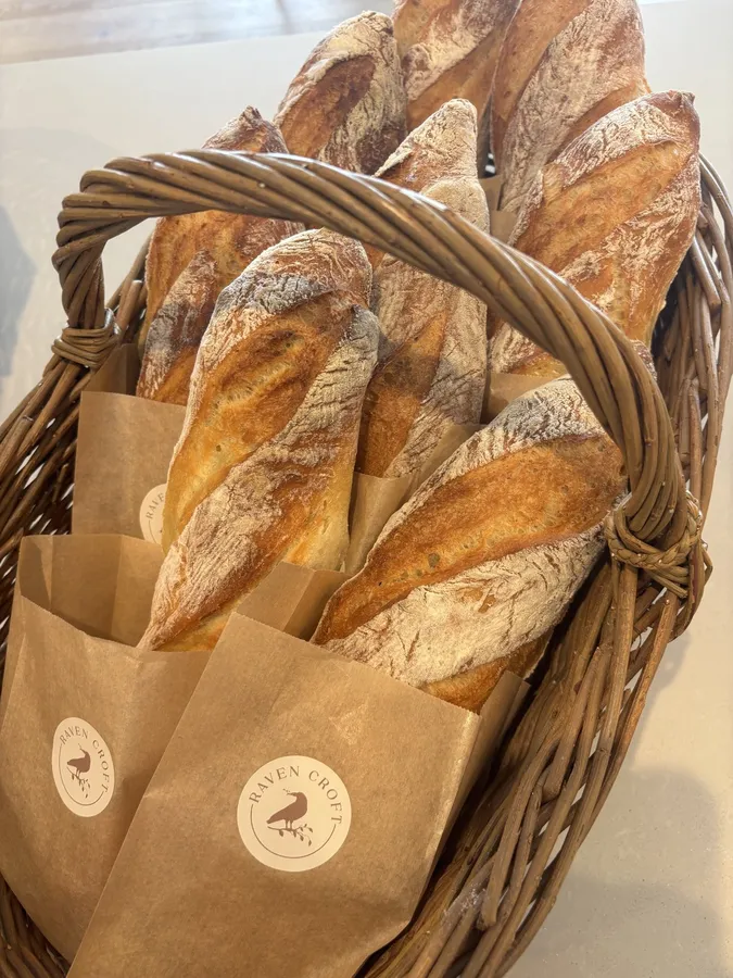A wicker basket packed with rustic baguettes in paper bags with Raven Croft stickers.