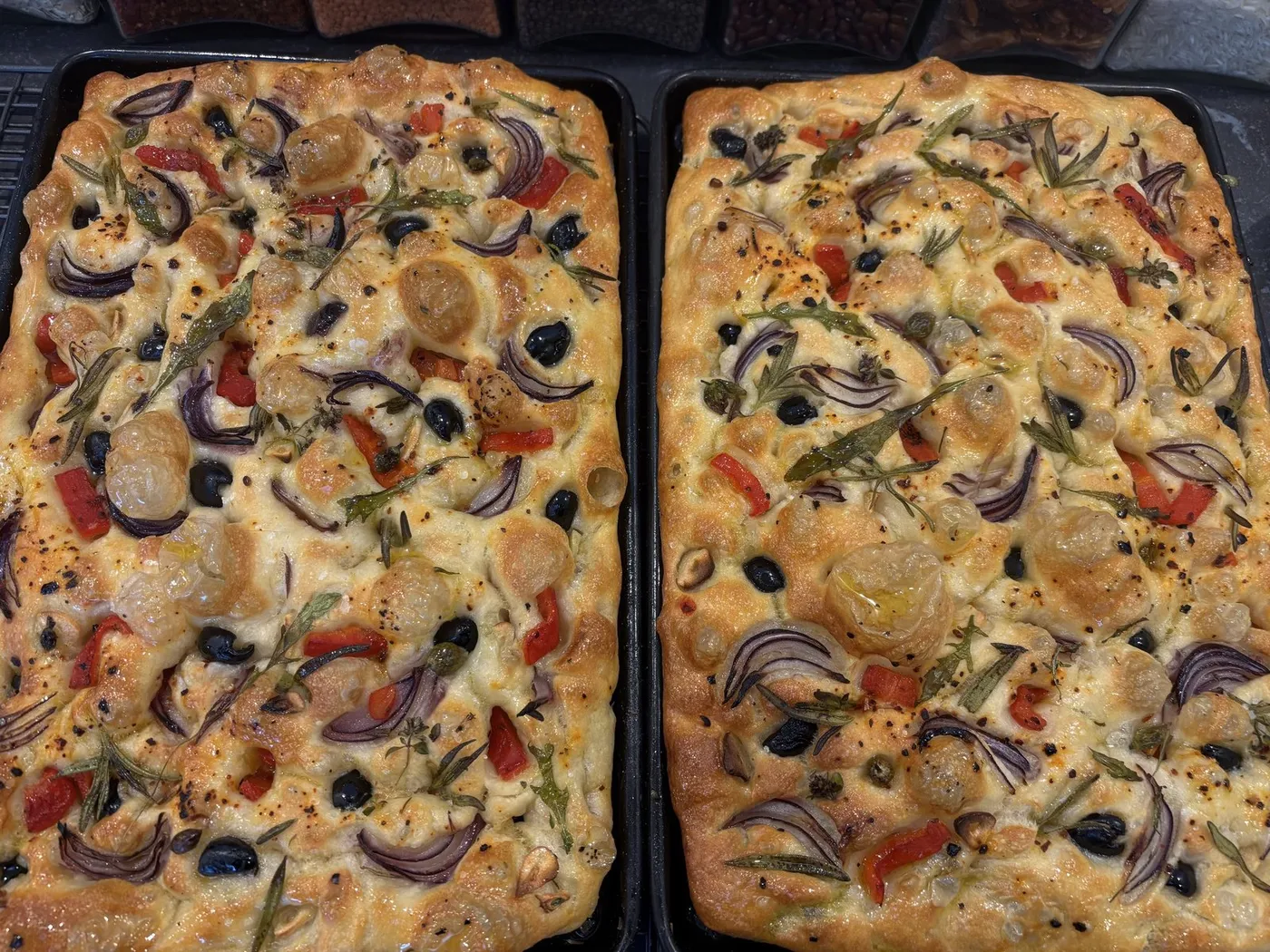 Two trays of focaccia topped with tomatoes, olives, red onion and herbs.